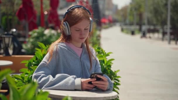 使用智能手机咖啡店的少女耳机在户外喝咖啡 坐在那里观看音乐电视的快乐的年轻女子 背对着花朵的背景音乐 — 图库视频影像