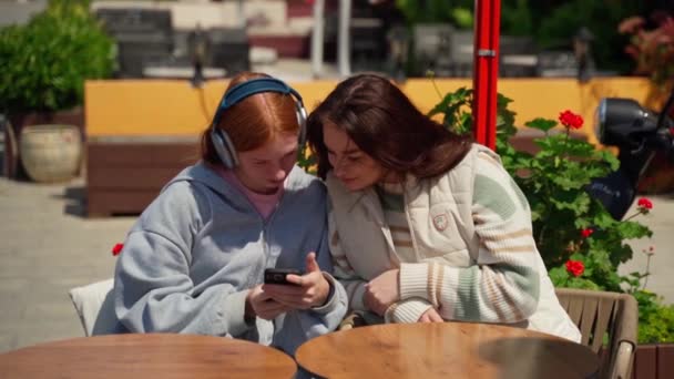 两个十几岁的女孩在咖啡馆里用智能手机观看视频照片内容 社交媒体在室外喝咖啡 使自己成为夏日阳光灿烂的自己 年轻女人说话 — 图库视频影像