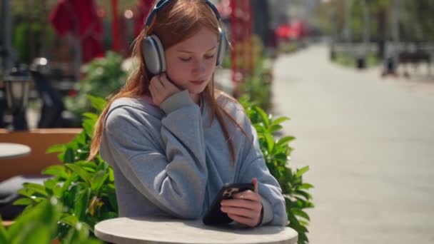 使用智能手机咖啡店的少女耳机在户外喝咖啡 坐在那里观看音乐电视的快乐的年轻女子 背对着花朵的背景音乐 — 图库视频影像