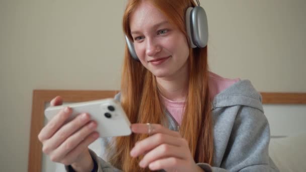 少女耳机在家里的床上用智能手机 年轻女人听音乐在线播客观看视频与小玩意 在线聊天聊天与朋友的互联网技术 — 图库视频影像