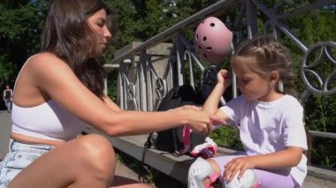 Anne yaz aylarında halka açık parkta kızına paten kayar dirsek koruması takıyor. Aile eğlencesi açık hava sporları oyunu.