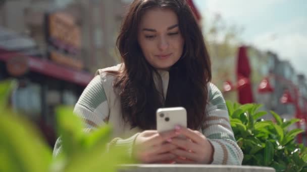 快乐的年轻女子在户外用手机打字 在城市背景下 快乐的少女坐在智能手机旁 社交媒体 暴民应用程序 观看视频 网上购物 浏览互联网 — 图库视频影像