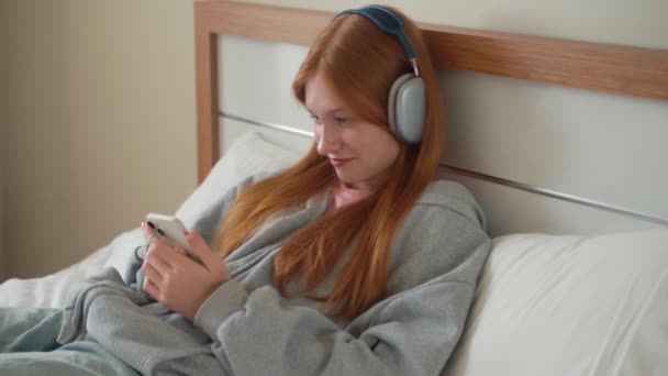 少女耳机在家里的床上用智能手机 年轻女人听音乐在线播客观看视频与小玩意 在线聊天聊天与朋友的互联网技术 — 图库视频影像