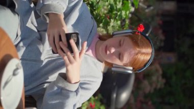 Genç kız kulaklıkları açık havada akıllı telefon kafesi kullanıyor kahve içiyor. Neşeli genç bir kadın oturmuş video izliyor çiçek arka planında müzik dinliyor dikey olarak.