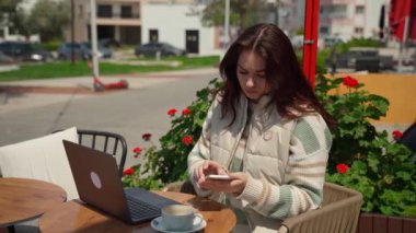 Kafede dizüstü bilgisayar kullanan genç bir kız kahve içiyor. Neşeli genç bir kadın oturmuş online öğrenci makalesi okuyor, sınav çalışması yapıyor çiçek arkaplanındaki akıllı telefonu konuşuyor.