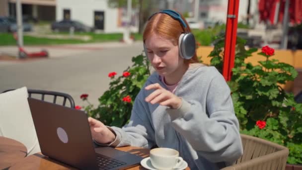 少女耳机在咖啡店外面喝咖啡时使用笔记本电脑智能手机 快乐的年轻女子坐在那里看电视 听着音乐电话 学生们写着作业 在花丛中学习考试 — 图库视频影像