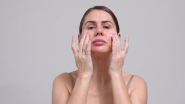 Orta yaşlı kadın, gri arka plan. 40 yaşında kadın nemlendirici krem suratı uyguluyor. Yüz masajı güzelliğinin, kendine olan saygısının ve sağlığının tadını çıkarıyor. Doğal Kozmetik Cilt Ürünleri.