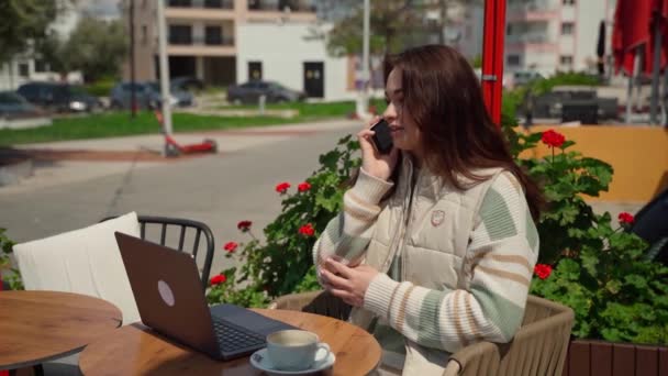 十几岁的女孩在咖啡馆里用笔记本电脑喝着咖啡 快乐的年轻女子坐在网上看学生的作文 在花朵的背景下说着聪明的电话 — 图库视频影像