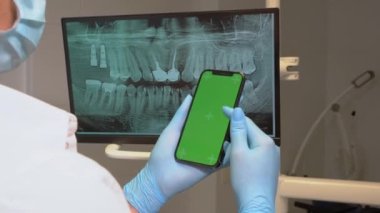 Kapalı diş hekimi steril eldiven, yeşil ekran cep telefonunu diş hekimliği ofisinde monitörde diş röntgeninin üzerinde tutuyor. Ortodontist kaydırma akıllı telefon kroma anahtar model şablonu dişler için görüntüde.