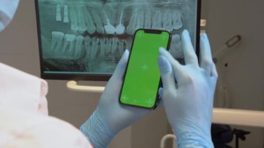 Kapalı diş hekimi steril eldiven, yeşil ekran cep telefonunu diş hekimliği ofisinde monitörde diş röntgeninin üzerinde tutuyor. Ortodontist kaydırma akıllı telefon kroma anahtar model şablonu dişler için görüntüde.