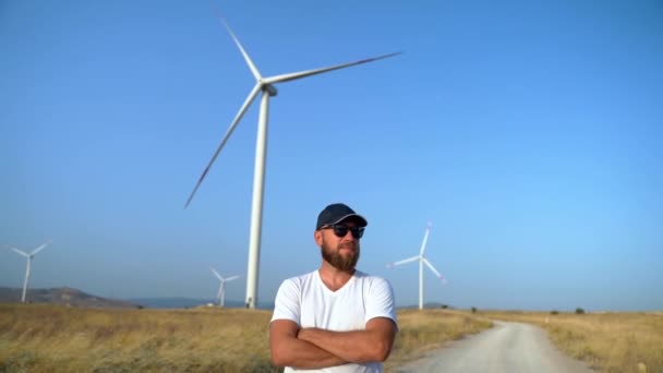 風車の風力発電機が付いている分野の田舎の道の上の箱の上に十字の腕に立っている幸せな自信のある男 環境に優しいエネルギーパワー 持続可能な消費 — ストック動画