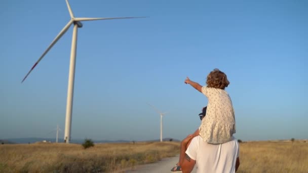 子供の少女を肩に乗せて風の農場を歩いている男の背中を撃った 幸せな父と娘 より環境に配慮した持続可能な空気を一緒に楽しむ — ストック動画