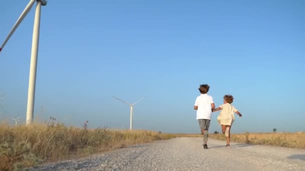 風車の風力発電機でフィールドで子供少女カントリーロードを実行している幸せなケアフリーの子供たち 子供時代 環境に優しいエネルギー — ストック動画