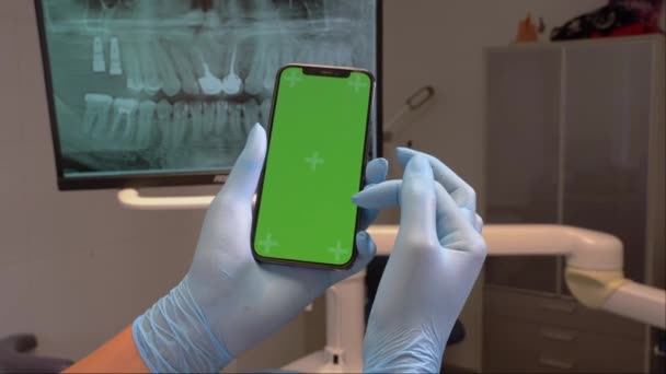 歯医者の手を閉じる滅菌手袋は モニター上の歯のX線の上に歯科事務所で緑のスクリーン携帯電話を保持します 歯医者は歯のためのディスプレイ上のスマートフォンのクロマキースクリーンモックアップテンプレートをスクロールします — ストック動画