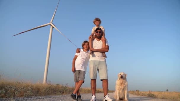 幸せな家族の父親の子供ゴールデンレトリーバーの犬の子供の少女は風車の風力発電機が付いている田舎道に立っています 子供時代 環境に優しいエネルギー — ストック動画