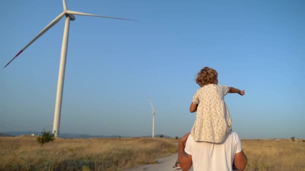 子供の少女を肩に乗せて風の農場を歩いている男の背中を撃った 幸せな父と娘 より環境に配慮した持続可能な空気を一緒に楽しむ — ストック動画