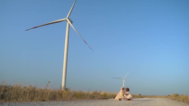 風車の風力発電機が付いている田舎の道の金の取り戻し犬が付いている幸せな世話のない子供の少女 子供時代 環境に優しいエネルギー — ストック動画