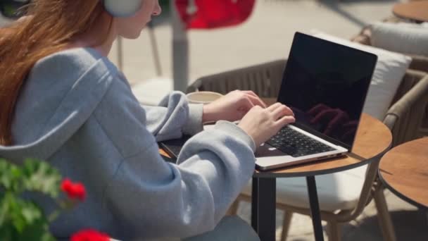 在咖啡馆里用笔记本电脑在户外喝咖啡的少女耳机 快乐的年轻女子坐在那里看视频 听着音乐电话学生的作业论文 在花卉背景色下的考试学习 — 图库视频影像