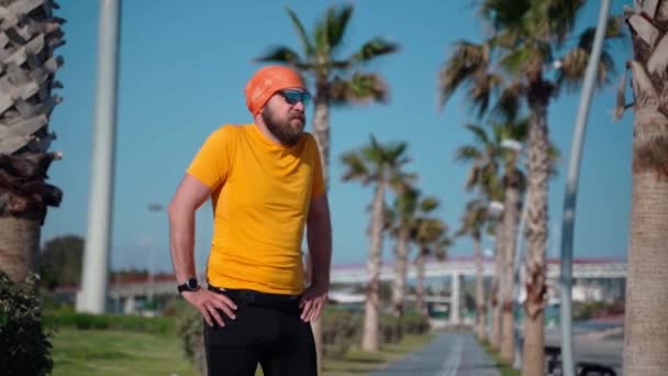 スポーツウェアのボディ正の男はジョギングマラソン晴れた日のトレッドミルヤシの背景のためのワークアウトの準備を実行する前に筋肉や関節のウォームアップを行います — ストック動画