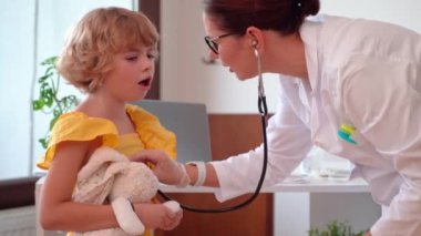 Klinikteki sağlık muayenesinde çocuğun ciğerlerini kontrol eden kadın doktor. Dost canlısı çocuk doktoru, steteskop kullanarak genç bir hastanın nefes alışını ve kalp atışlarını inceliyor.