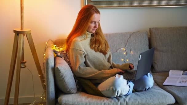 红头发的年轻女人坐在沙发舒适的家使用笔记本电脑 圣诞节期间穿上最喜欢的毛衣在家里工作 自由职业者在网上购物过冬 准备下订单发送电子邮件 — 图库视频影像