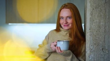 Kızıl saçlı genç kadın evde rahat, sıcak ve sıcak bir süveter giyiyor. Anne, bebek uyurken şifalı bitkilerle sıcak çay içiyor. Samimi bir rahatlama ve sükunet anı. Gerçek anın tadını çıkarıyorum..