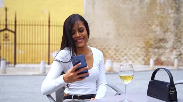 漂亮的拉丁裔美国女人坐在咖啡厅的庭院里用电话 滚动社交媒体 使用移动应用程序 订购在线菜单 与朋友聊天 创建内容 乘坐E — 图库视频影像
