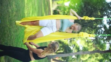 Aero hava yogası antrenörü eğitmeni hamağın kadın katılımcılarına Asana 'ya nasıl egzersiz güvenli ve sağlıklı yapılacağını anlatıyor ve yardımcı oluyor. Parkland 'da temiz hava alın. Kadınlar için esneme sporu