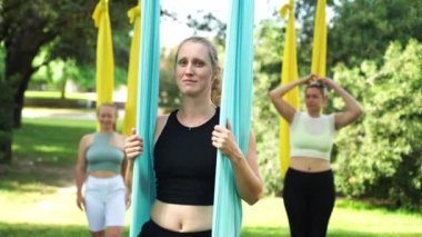 Kadın deneyimli yoga eğitmeni hamakta oturur ve kameranın orta portresine bakar. Parkın dışındaki spor salonunda. Yetişkinlerin bakımı için grupta birlikte eğitim gören kadınlar