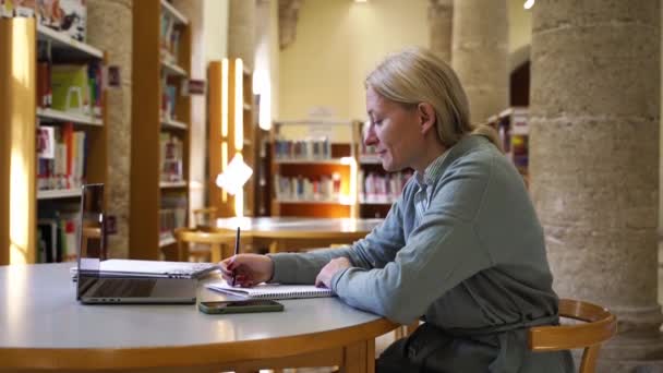 在图书馆学习新教育科目的成年女生中年妇女在网上使用笔记本电脑学习和研究科学资料 提高专业资格 — 图库视频影像