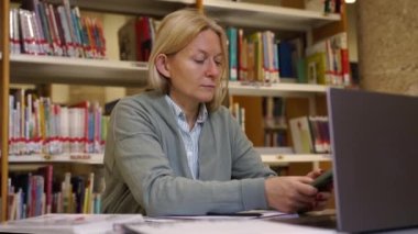 İş yerinde çalışan orta yaşlı bir kadın kütüphane kitaplarının arka planında dizüstü bilgisayar ve akıllı telefon kullanıyor. Bayan yetişkin öğretmen okul dersleri, bilimsel tez, materyal araştırmaları için hazırlanıyor..