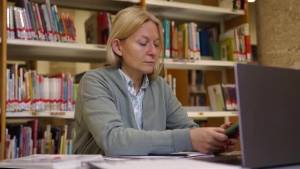 在同事办公室工作的中年妇女 在图书馆图书的背景下使用笔记本电脑和智能手机 成年女教师准备学校课程 科学论文 材料研究 — 图库视频影像