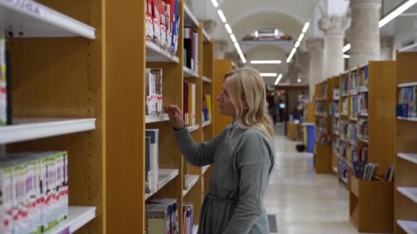 図書館や書店で本を選択する中年女性 大人は本を通して飛び散る 本屋で読書する学生 文学のような成熟した女性 図書館で本を選ぶ — ストック動画