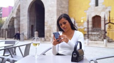 Kafe avlusunda telefon kullanan güzel Latin Amerikalı kadın. Sosyal medya kaydırma, mobil uygulama kullanma, çevrimiçi menü siparişi verme, arkadaşlarla sohbet etme, içerik oluşturma, makara oluşturma, e