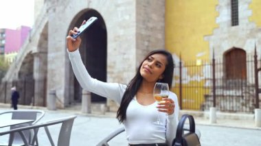 Halka açık veranda veranda restoranında akıllı telefon kullanan Latin kökenli genç bir kadın. Arkadaşlarla video görüşmesi, sosyal medya için içerik yaratmak, Selfie. Şarap içmek