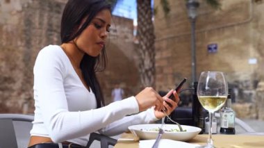 Kafe avlusunda telefon kullanan güzel Latin Amerikalı kadın. Sosyal medya kaydırma, mobil uygulama kullanma, çevrimiçi menü siparişi verme, arkadaşlarla sohbet etme, içerik oluşturma, makara oluşturma, e