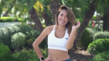 Asyalı yetişkin kadın koşucunun yakın plan yüzü spor kıyafetleri içinde yeşil orman arka planında kameraya bakıp gülümsüyor güneşli bir sabah çalıştırmadan önce Kentsel sağlıklı yaşam tarzı