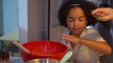 Siyah tenli çocuk krep pişiriyor, hamuru sıcak mutfakta kaşıkla kaseye karıştırıyor. Çok ırklı ailelerin öğrettiği küçük melez kız modern mutfakta sağlıklı yemekler pişiriyor..