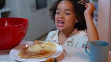 Afro-Amerikalı komik kız evdeki rahat mutfakta krep pişirirken krep yiyor. Çok ırklı aile geleneği hafta sonu tatili