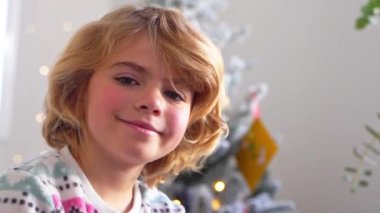 Gülümseyen ilkokul çocuğunun portresi Noel ağacı süsleme arkaplanından kameraya bakıyor, reklam için negatif alanı kopyalıyor Noel Baba 'ya yeni yıl tatili için mektup yazıyor, rahat bir ev.