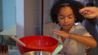 Siyah tenli çocuk krep pişiriyor, hamuru sıcak mutfakta kaşıkla kaseye karıştırıyor. Çok ırklı ailelerin öğrettiği küçük melez kız modern mutfakta sağlıklı yemekler pişiriyor..