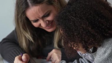Kafkasyalı anne ve Afrikalı Amerikalı kız evde çalışıyor, rahat beyaz odada ödev yapıyor. Ebeveyn öğretmenliği, çocuk okulu kızı bilgi öğrenme, egzersiz kitabı yazma. Çocuklar için eğitim
