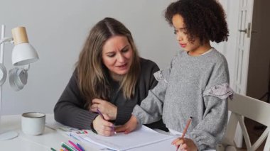 Kafkasyalı anne ve Afrikalı Amerikalı kız evde çalışıyor, rahat beyaz odada ödev yapıyor. Ebeveyn öğretmenliği, çocuk okulu kızı bilgi öğrenme, egzersiz kitabı yazma. Çocuklar için eğitim