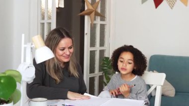 Kafkasyalı anne ve Afrikalı Amerikalı kız evde çalışıyor, rahat beyaz odada ödev yapıyor. Ebeveyn öğretmenliği, çocuk okulu kızı bilgi öğrenme, egzersiz kitabı yazma. Çocuklar için eğitim