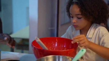 Afro-Amerikalı çocuk krep pişiriyor, hamuru sıcak mutfakta kaşıkla bir kaseye karıştırıyor. Küçük melez kızımız modern mutfak akşamında sağlıklı yemekler pişiriyor..