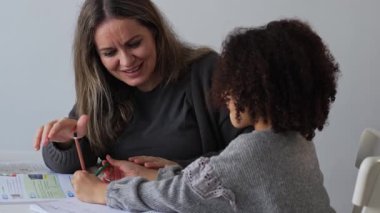 Kafkasyalı anne ve Afrikalı Amerikalı kız evde çalışıyor, rahat beyaz odada ödev yapıyor. Ebeveyn öğretmenliği, çocuk okulu kızı bilgi öğrenme, egzersiz kitabı yazma. Çocuklar için eğitim