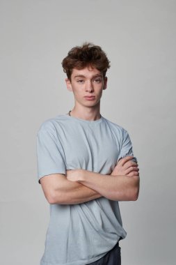 Bir fotoğraf stüdyosunda dururken kolları çaprazlanmış kameraya bakan tişörtlü bir çocuğun portresi.