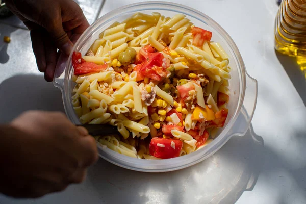 From above, an anonymous person preparing a portion of tasty pasta with tuna, vegetables and olives in a plastic bowl on a sunny day