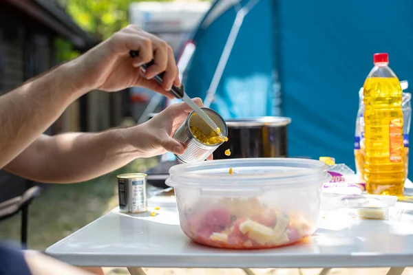 Unrecognizable male adding canned corn into plastic bowl with salad while sitting at table and cooking lunch on campsite
