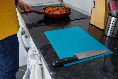 Anonymous person moving a frying pan and next to it a cutting board with a knife